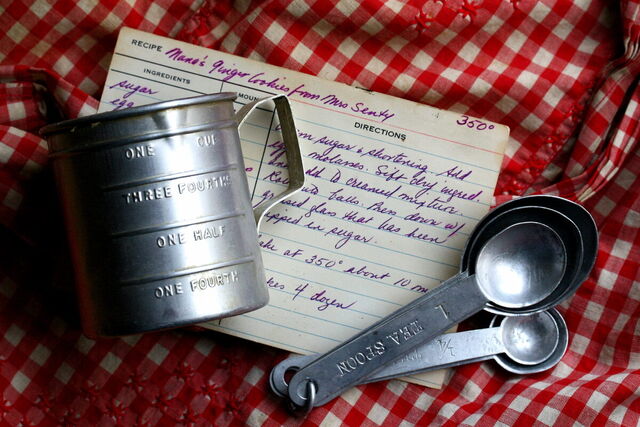 A handwritten recipe card with a flour sifter and measuring spoons on a gingham cloth